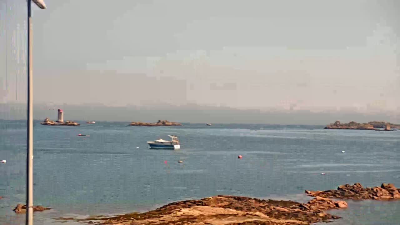 A hazy view of a calm sea shows small islands, a boat, and rocky shoreline in the foreground.