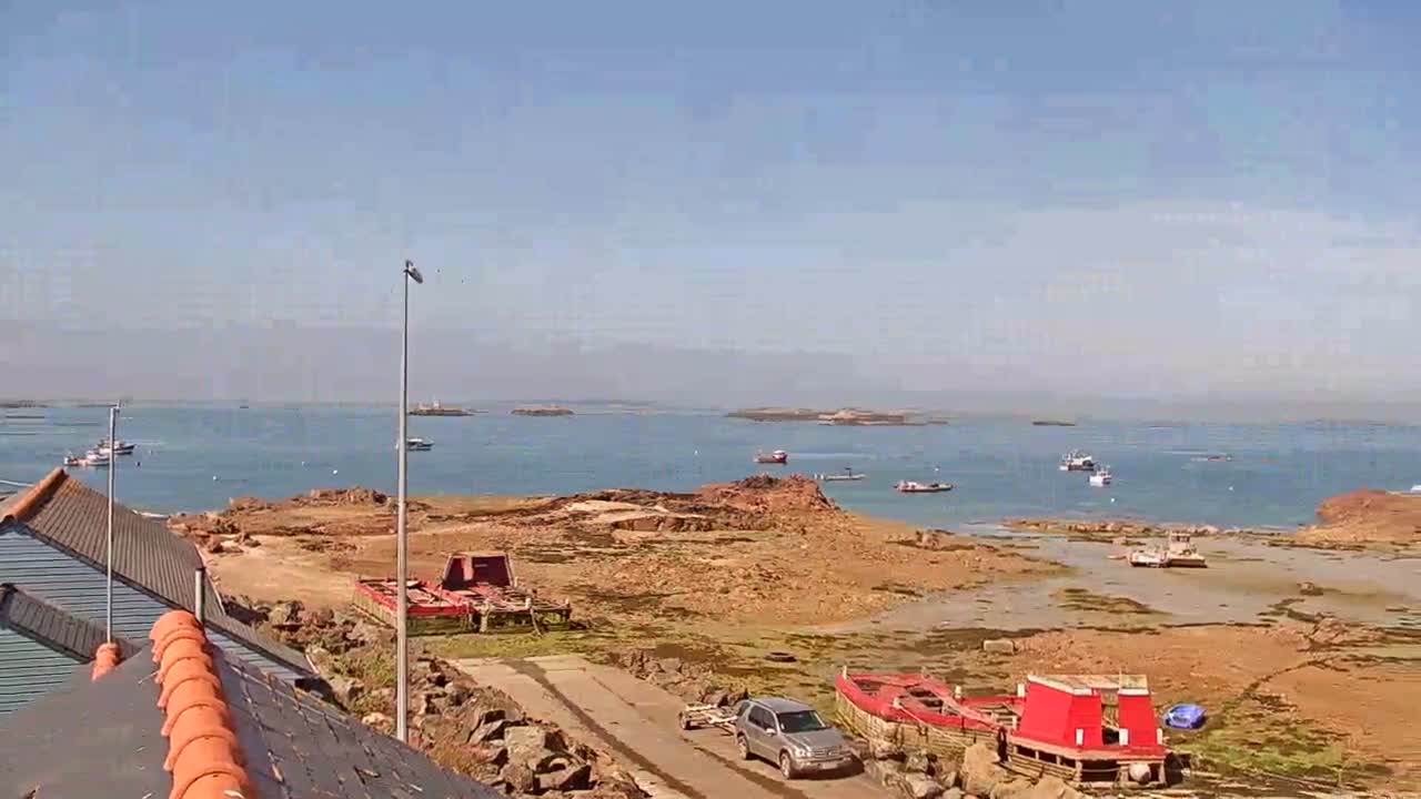 A sunny day reveals a rocky shoreline with several boats, both in the water and beached, and a vehicle parked near a building with a red-tiled roof.