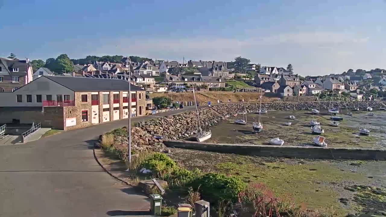 A small harbor with numerous sailboats resting on exposed mudflats under a clear blue sky, is situated next to a town of small houses and a large building.