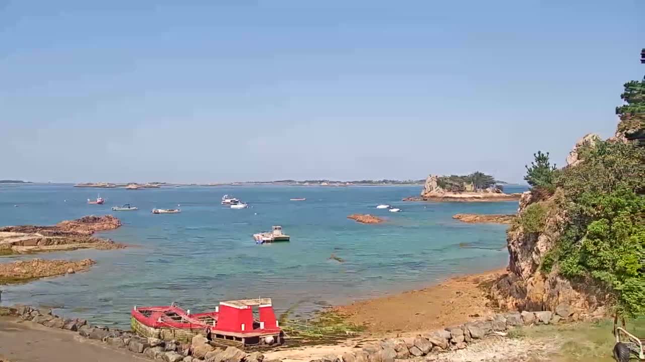 A calm, sunny day reveals a coastal scene with several small boats scattered across clear, shallow water near rocky shores and small islands.