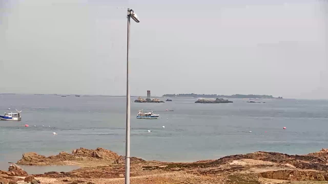 A calm, overcast day reveals a coastal scene with rocky shoreline, several small boats on the water, and distant islands.
