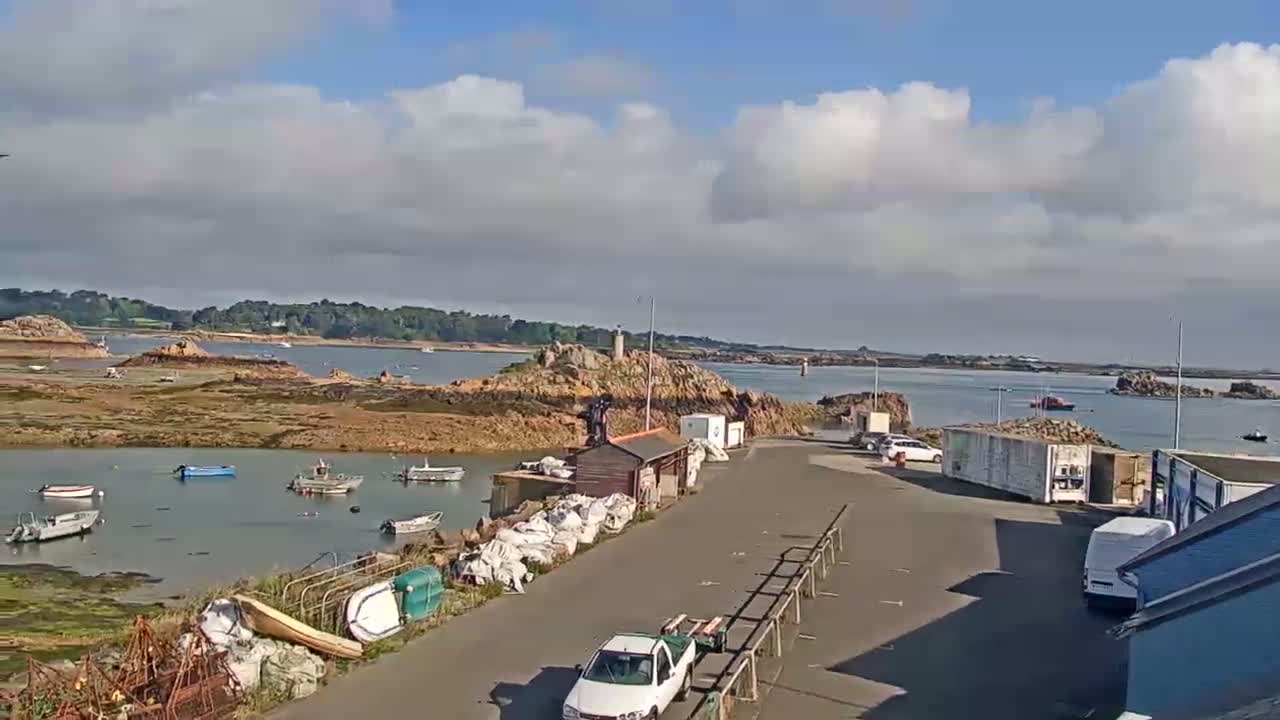 A mostly sunny day shows a harbor with several small boats, a rocky shoreline, and cars parked near a small building.