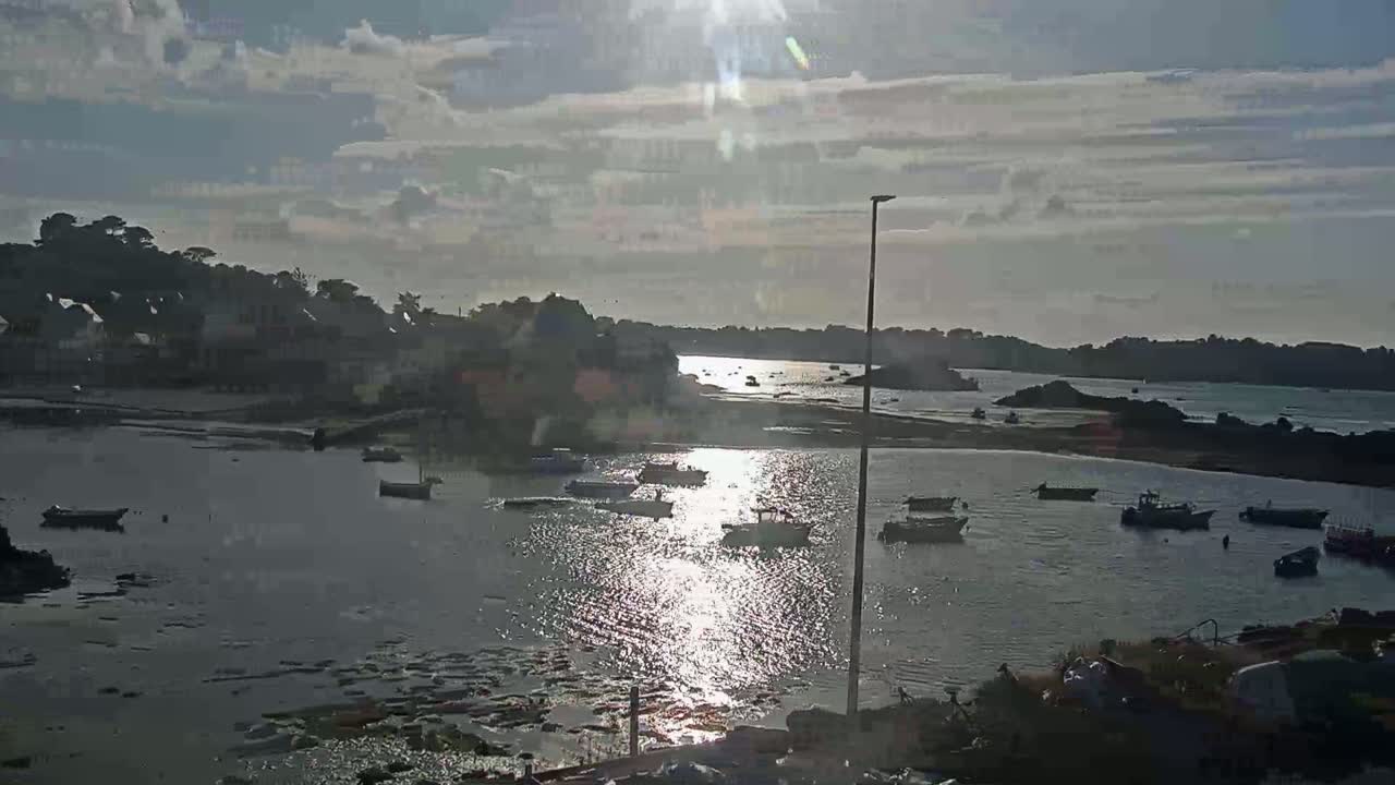 A calm bay with numerous small boats, a village in the background, and the sun shining brightly through partly cloudy skies.