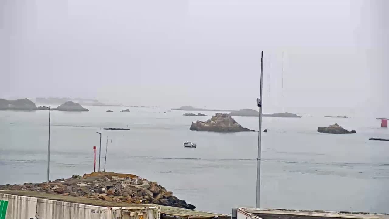 A foggy harbor scene shows several small rocky islands in calm water with a small boat and some harbor structures.
