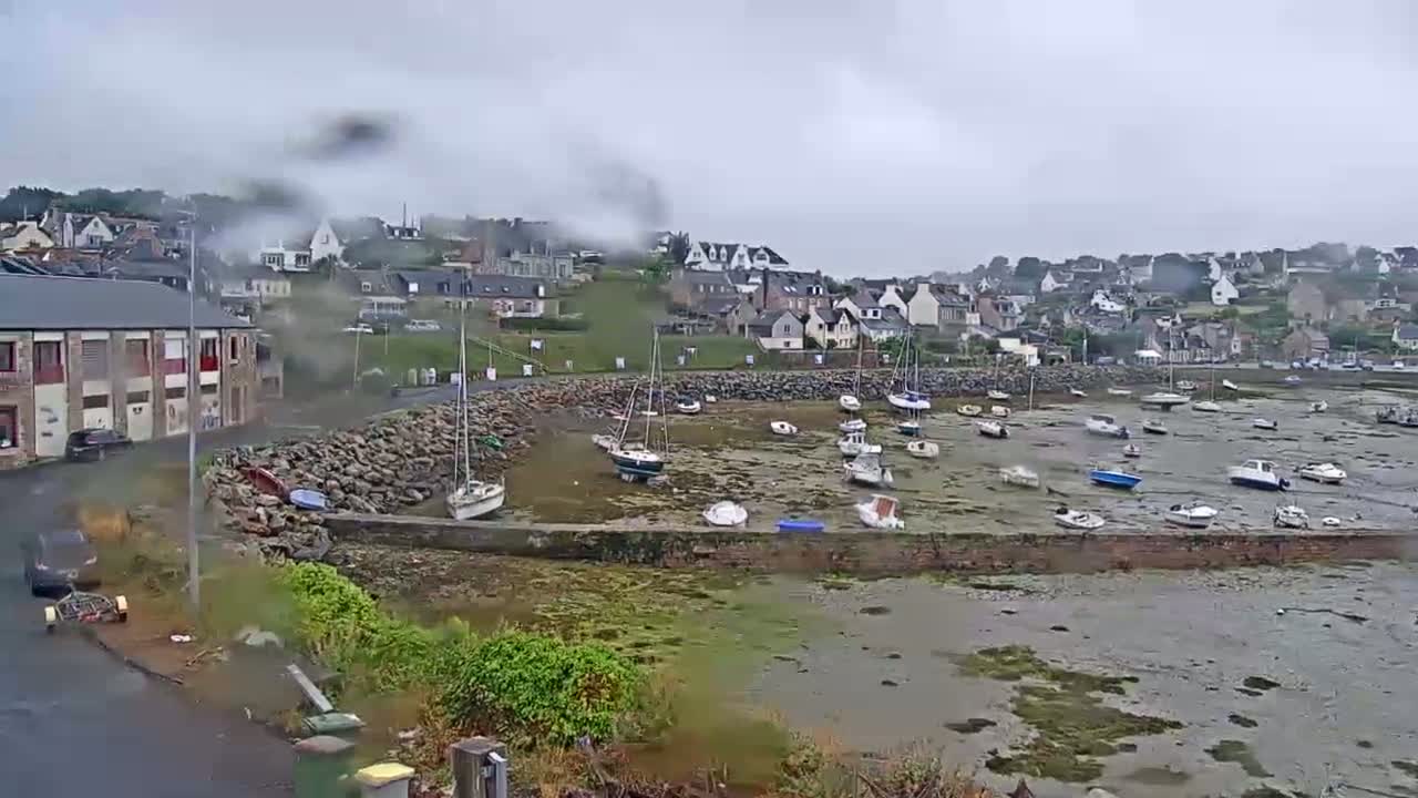 A small harbor filled with numerous boats sits at low tide, next to a small town under an overcast sky.