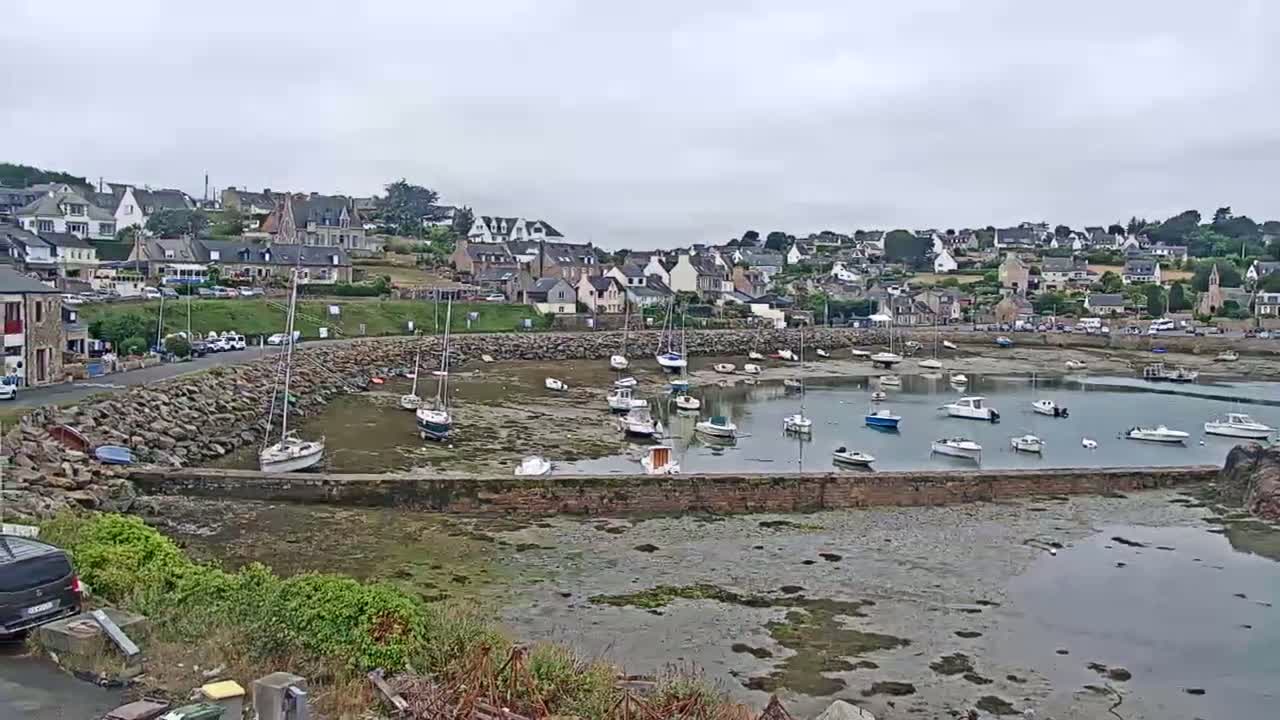 A small harbor filled with boats at low tide is nestled beside a village under an overcast sky.
