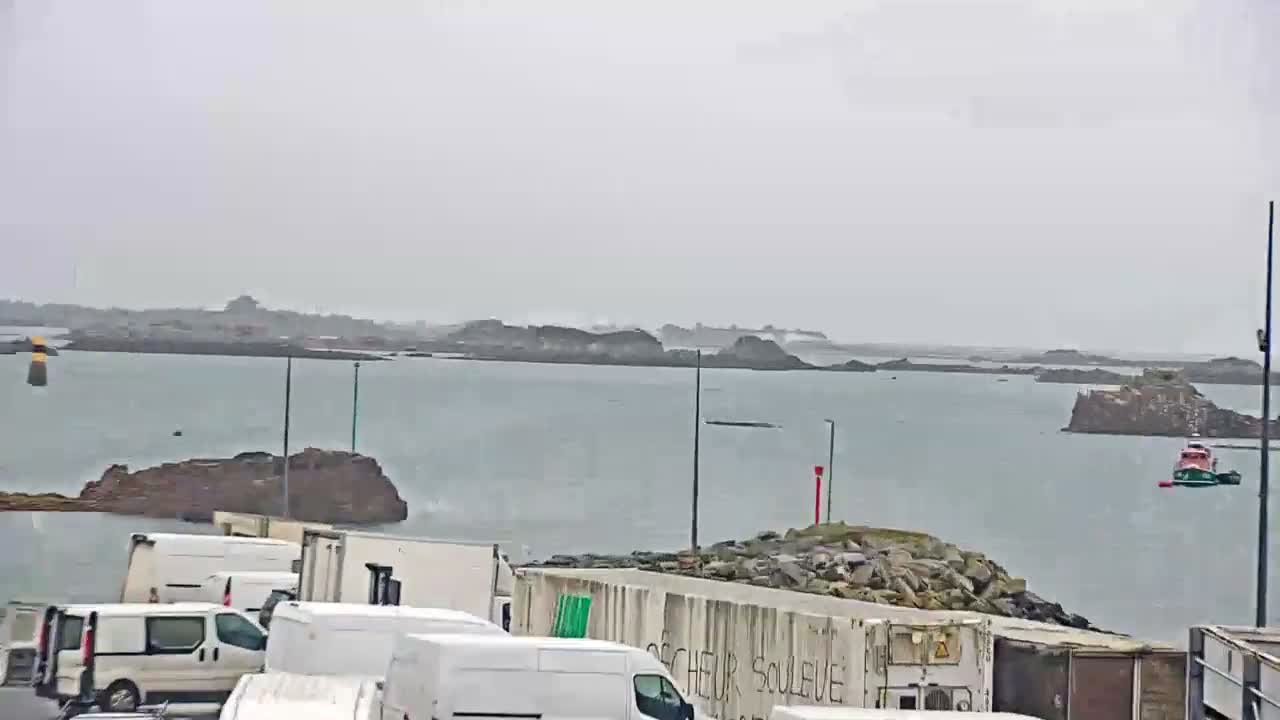 An overcast day casts a subdued light over a coastal scene featuring a bay dotted with numerous rocky islands and navigational markers, with a red and green boat on the water and white delivery vans parked along a rocky foreground embankment.