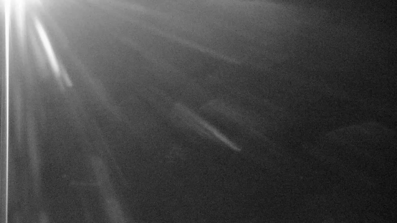 A monochromatic image depicts strong light flares and rays fanning from the upper left against a dark, grainy background, suggesting a hazy or dusty outdoor night.