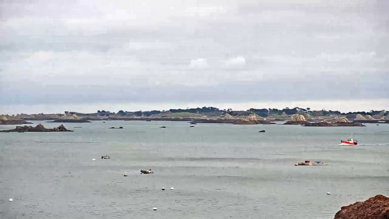 Under a cloudy, overcast sky, a grey sea is dotted with numerous rocky islets and a distant green coastline, while a red and white boat moves across the water, accompanied by a barge and several buoys, viewed from a rocky foreground.