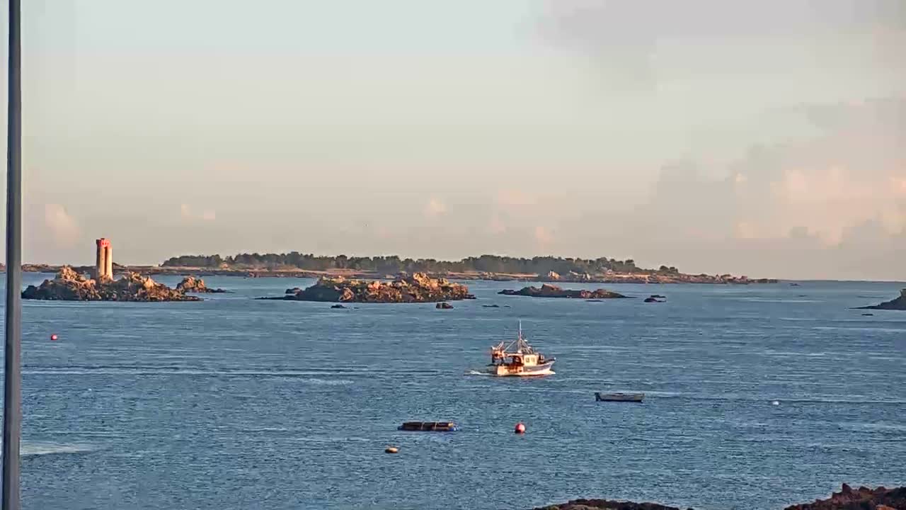 A fishing boat motors across calm blue waters, passing numerous rocky islets including one with a red and white lighthouse, under a bright, partly cloudy sky.