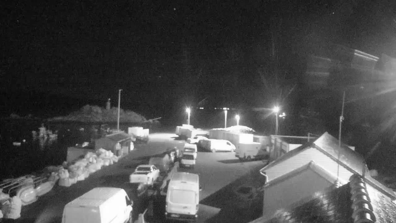 A clear night reveals a coastal scene illuminated by artificial lights, featuring numerous parked vehicles, buildings, and a distant lighthouse overlooking the water.