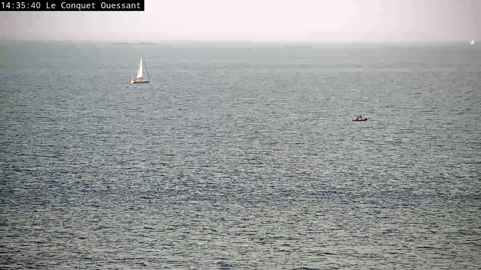 A sailboat and a small motorboat are visible on a calm, hazy sea.