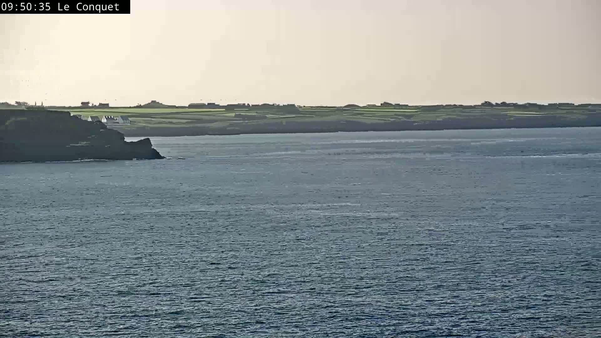 A calm body of water stretches out before a dark cliff face, behind which sits a line of houses and green pastureland under a hazy sky.