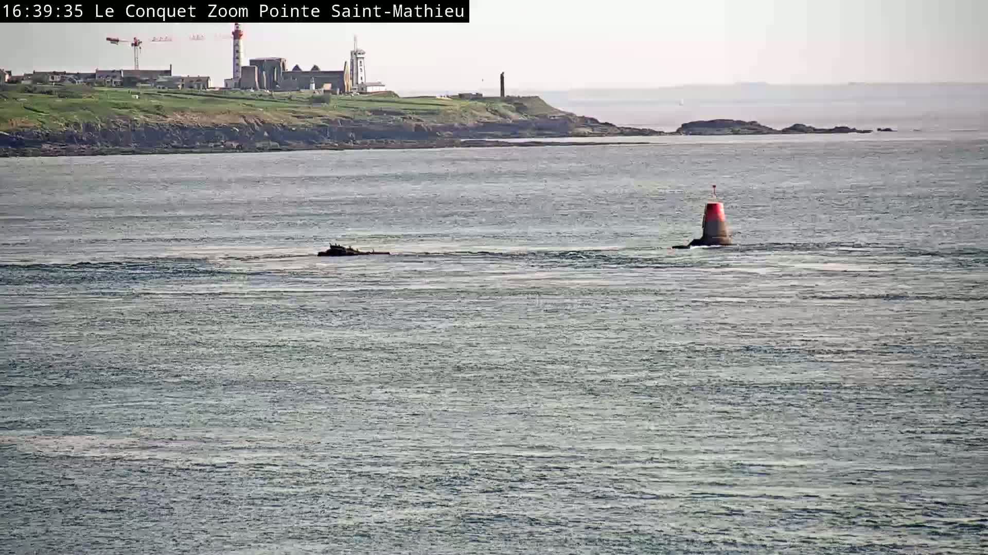 A calm body of water shows a red and gray navigational buoy and distant land with a lighthouse under a hazy sky.