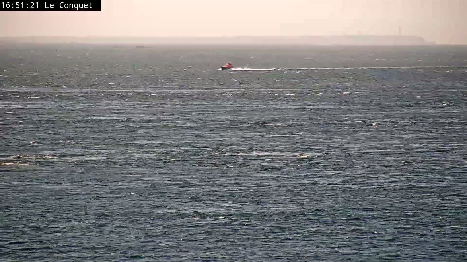 A small, red boat travels across a choppy, gray sea under a hazy sky.