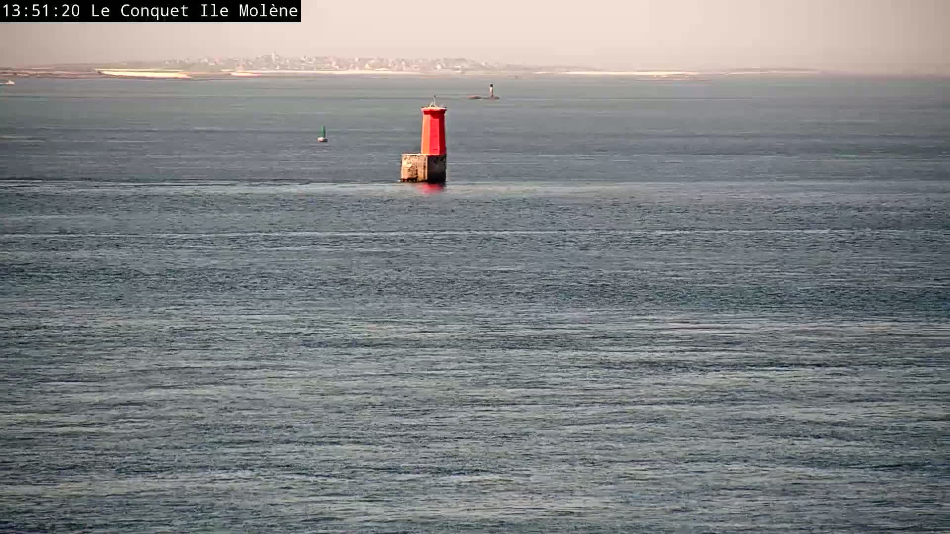 A red lighthouse stands in calm, gray-blue water under a hazy sky, with land barely visible in the far distance.