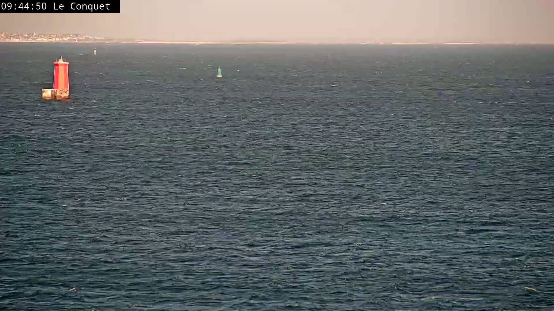 A red and white lighthouse sits in a dark-blue sea with a light chop under a hazy sky.