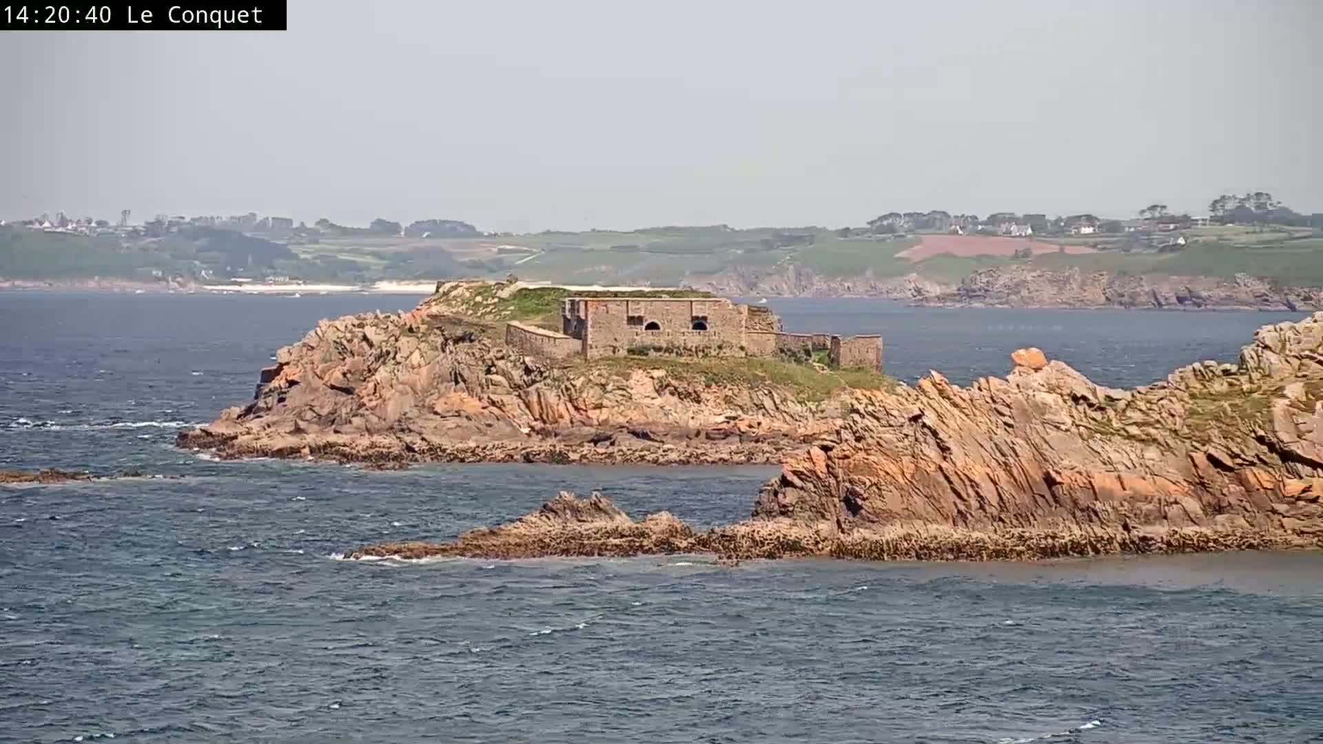 A stone fort sits on a rocky island in a calm sea, with a distant coastline visible under a hazy sky.