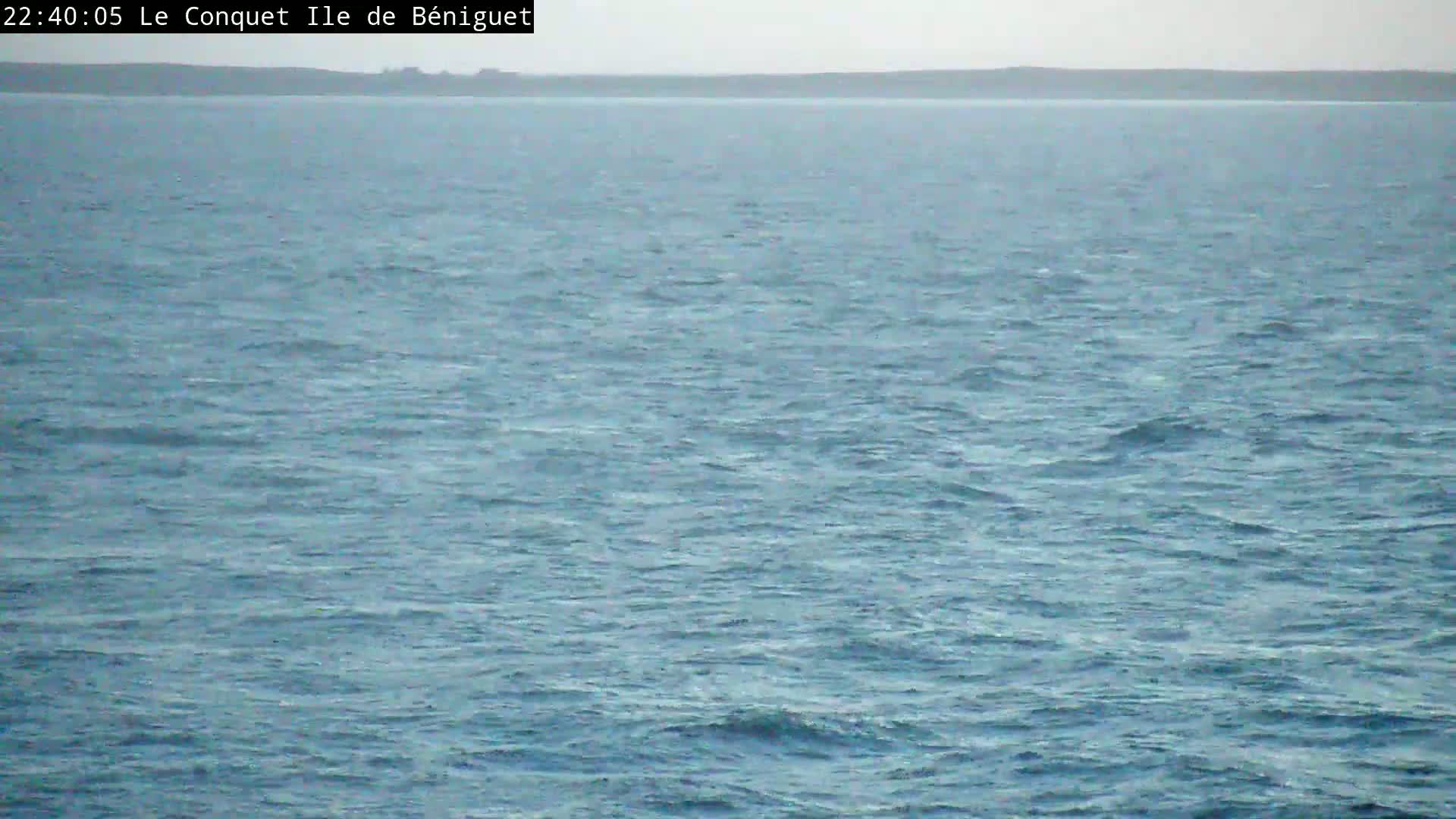A calm sea with small waves stretches to a distant, dark shoreline under an overcast sky.