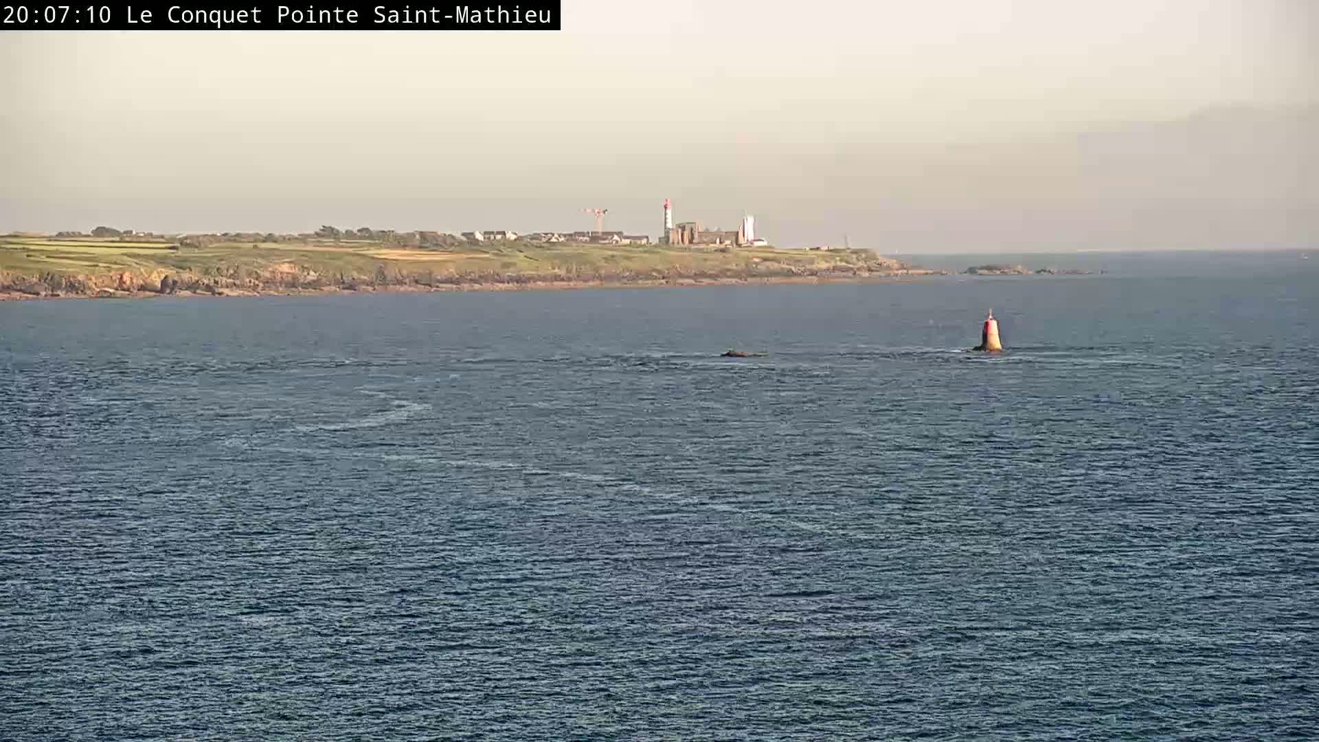 A calm body of water shows a distant coastline with a lighthouse and a smaller, red and white buoy closer to the viewer.
