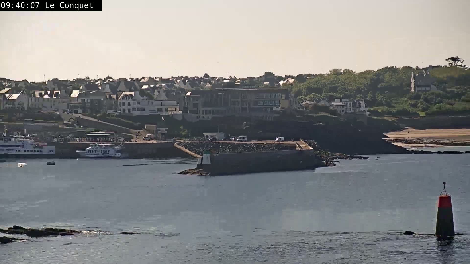 A coastal town with buildings on a hill overlooking a harbor with boats, a breakwater, and calm water under a mostly clear sky.
