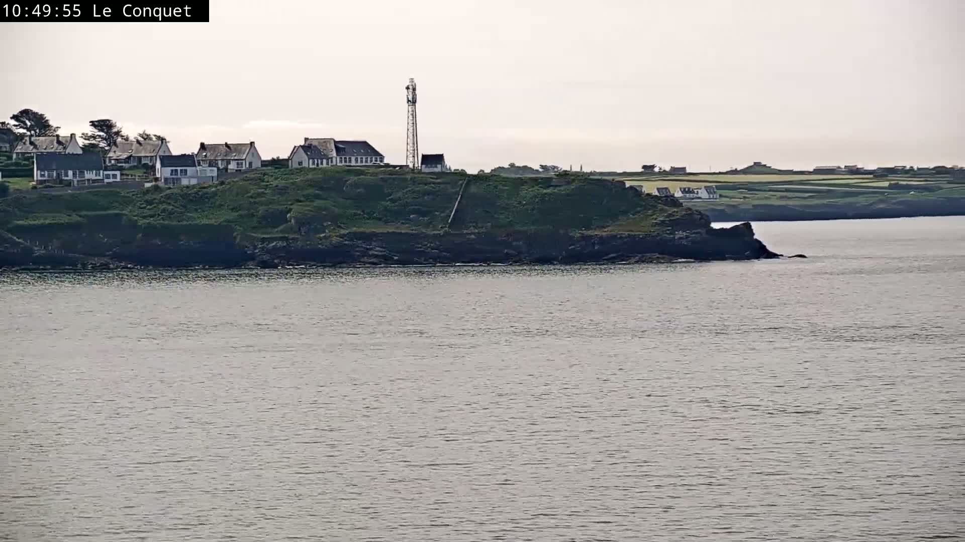 A coastal scene shows a cluster of houses atop a green, rocky cliff overlooking a calm body of water under an overcast sky.