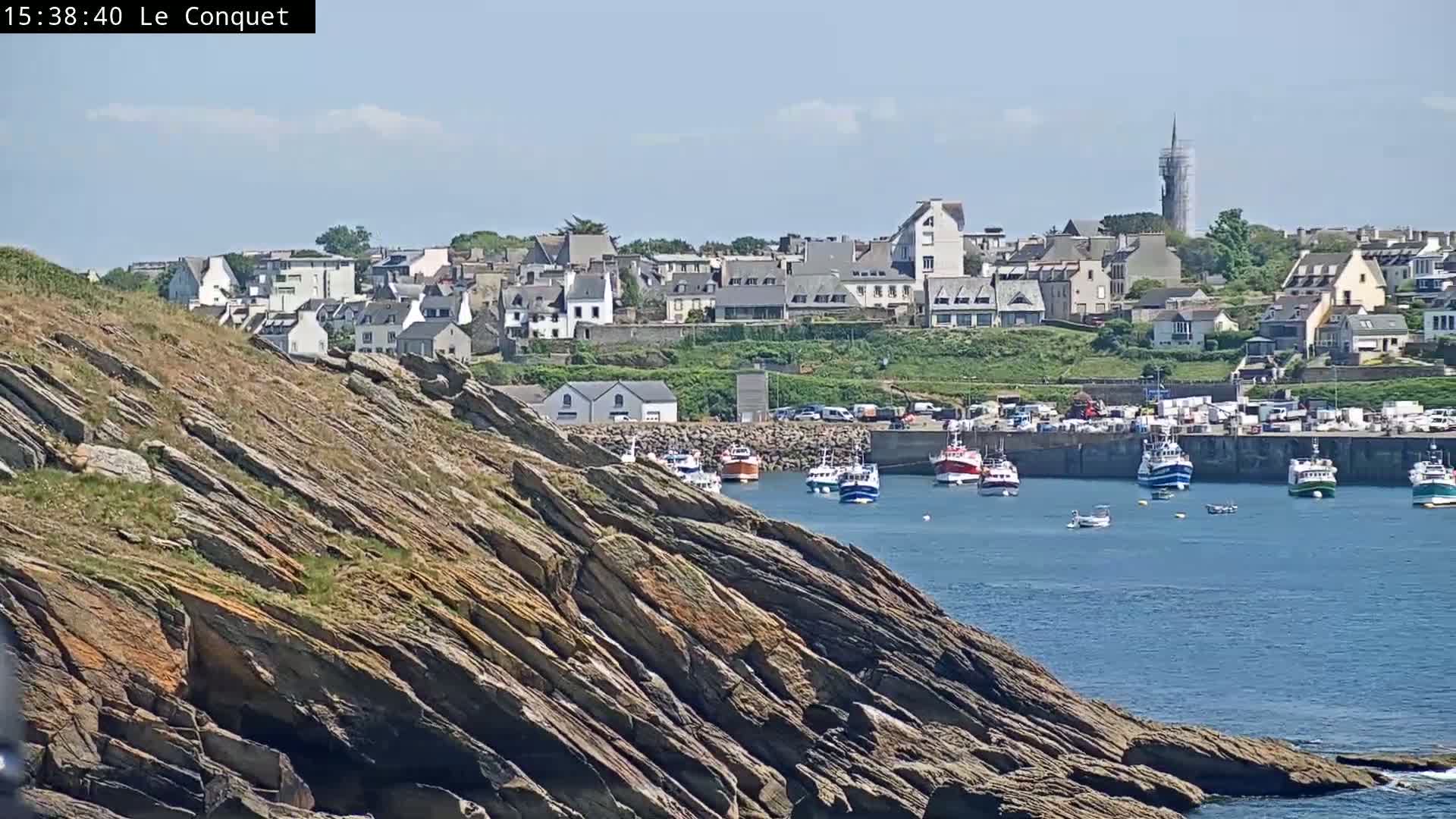 A rocky cliff overlooks a harbor filled with fishing boats, with a town visible in the background under a mostly sunny sky.