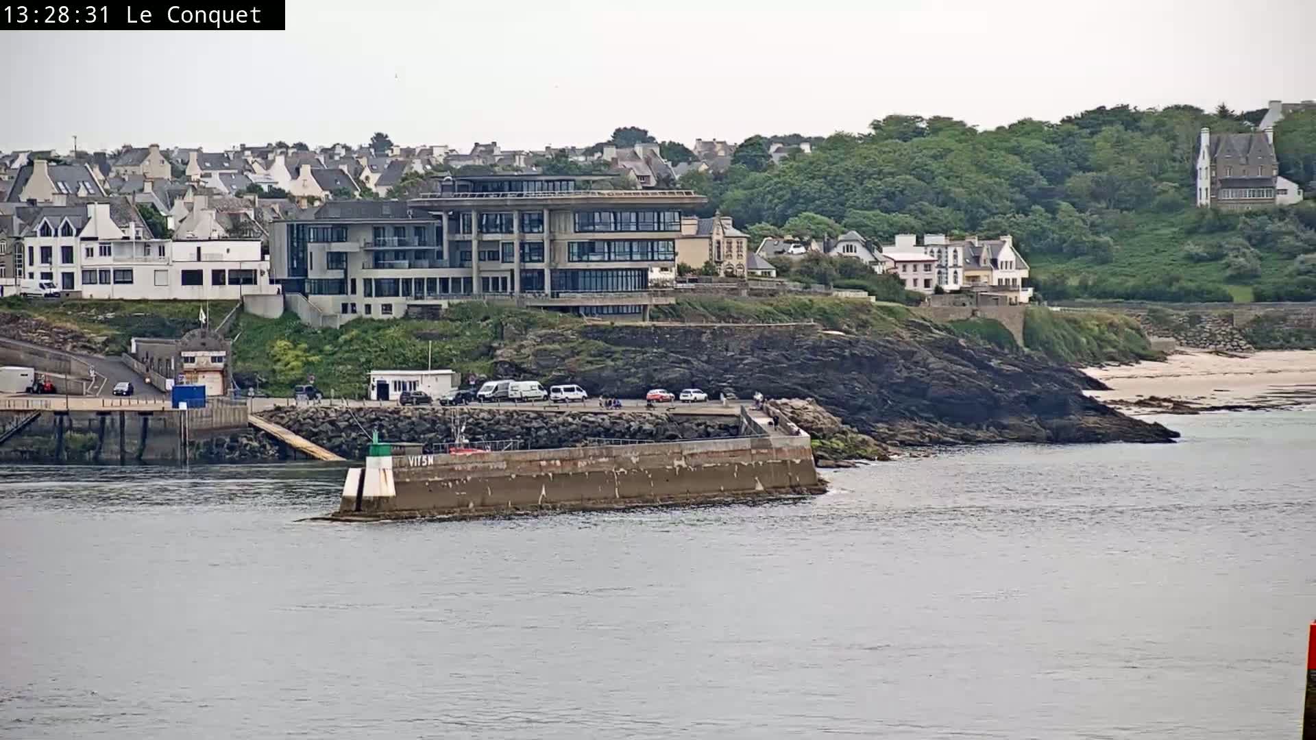 A coastal town with several buildings is partially visible from across the water on an overcast day, with a small breakwater and a few vehicles near the water's edge.