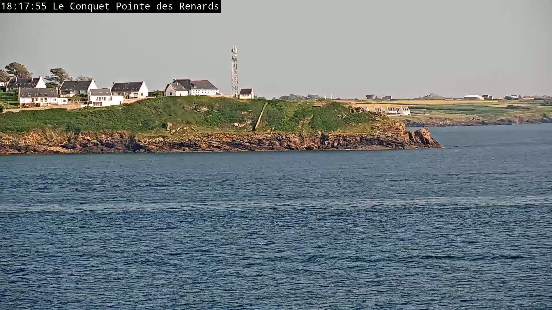 A rocky coast with several houses and a communications tower overlooks calm, dark-blue water under a clear, light-grey sky.