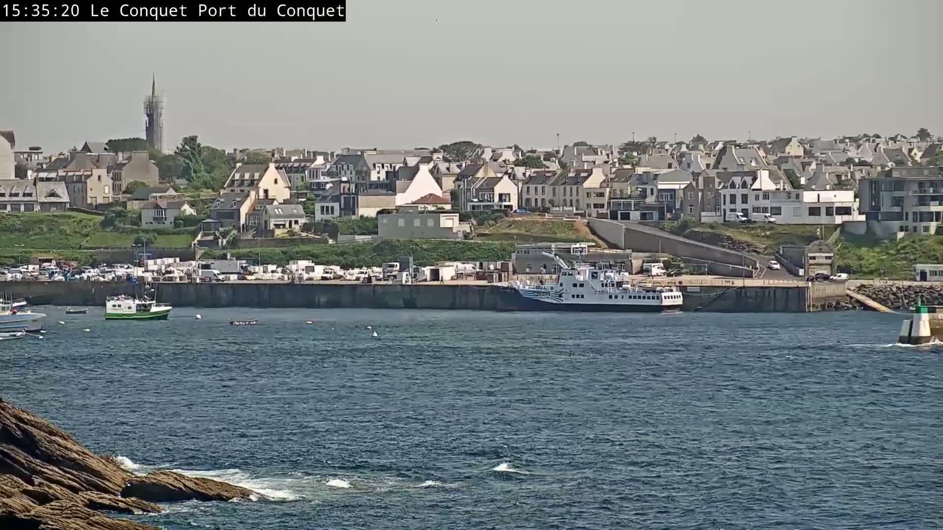 A ferry is docked at a harbor next to a town with a tall steeple visible in the distance, under a mostly clear sky.