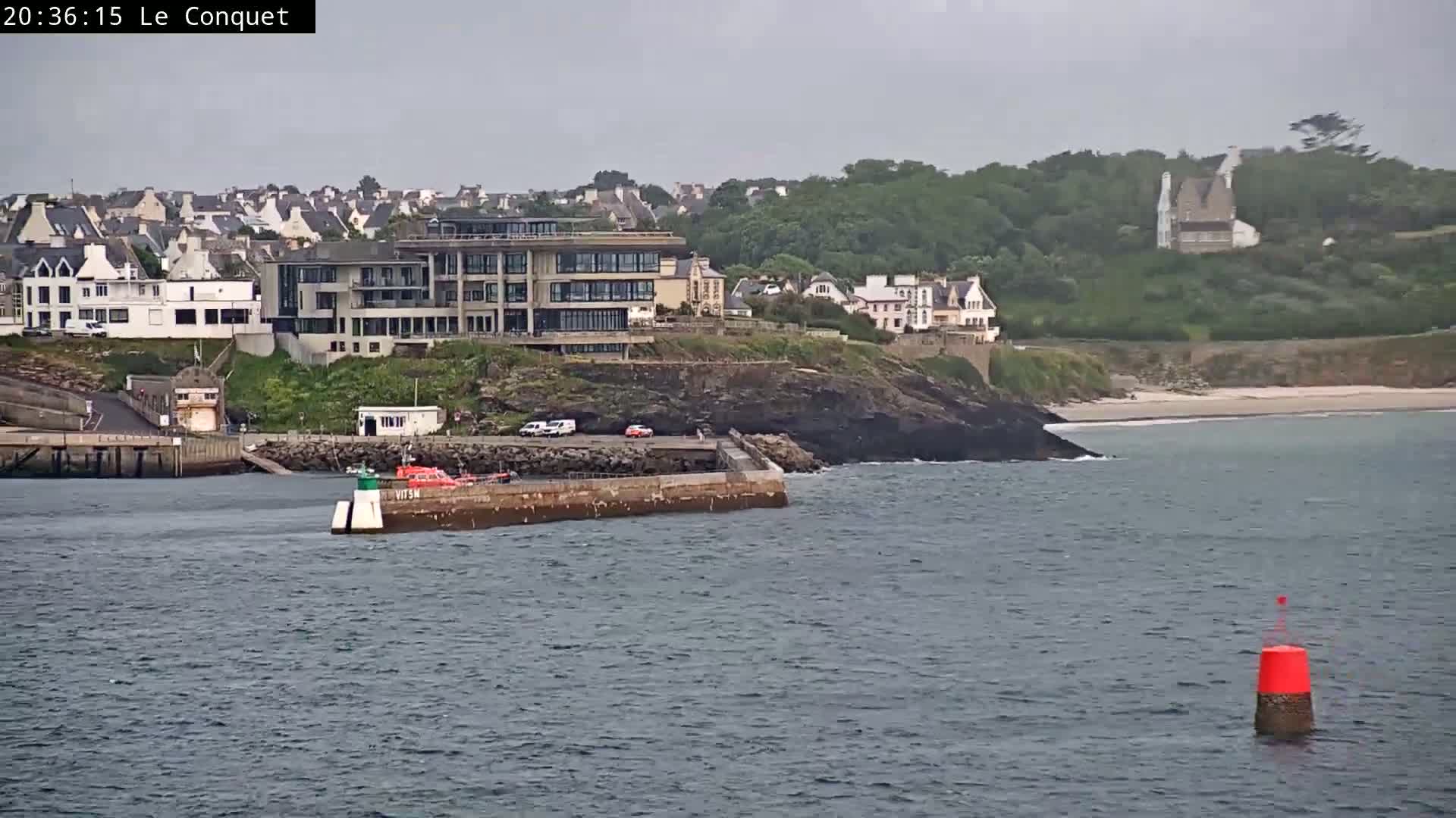 A coastal town with buildings on a cliff overlooking a harbor with a small boat, under an overcast sky.