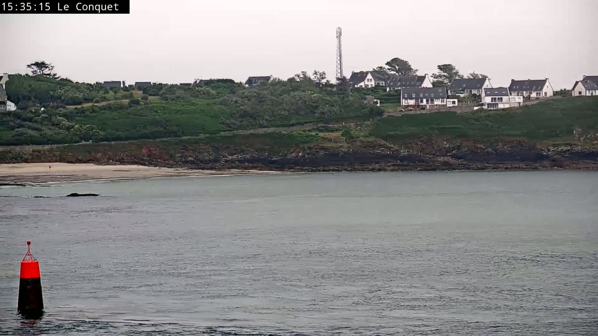 A calm body of water shows a small sandy beach and a line of houses on a green hill under an overcast sky.