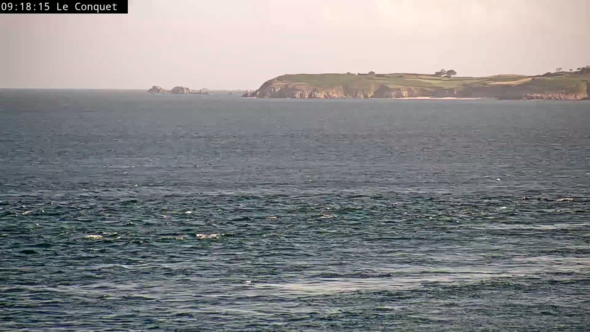 A moderately wavy ocean stretches to a distant, verdant, hilly coastline under a hazy sky.