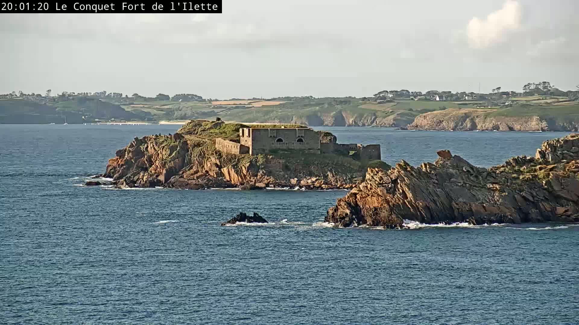 A stone fortress sits on a small, rocky island in a calm sea, with green hills visible in the background under a mostly clear sky.