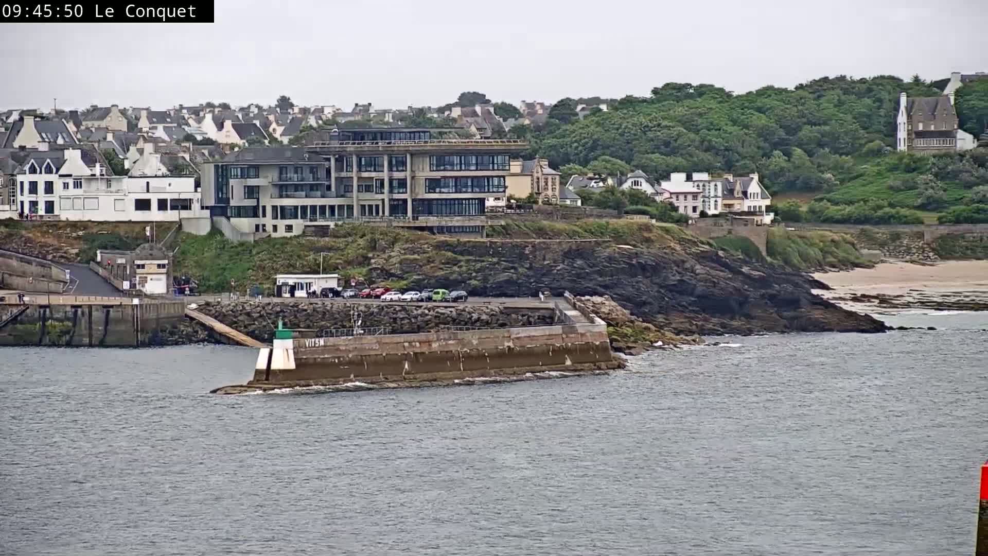 A coastal scene on an overcast day shows a modern building on a rocky cliff overlooking a small harbor with a breakwater and a parking lot, next to a town of white buildings and green hills.