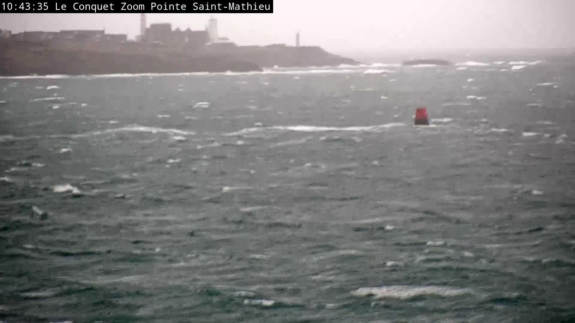A choppy, gray sea with a red buoy in the mid-ground stretches towards a hazy coastline featuring a lighthouse and other buildings under overcast, stormy conditions.