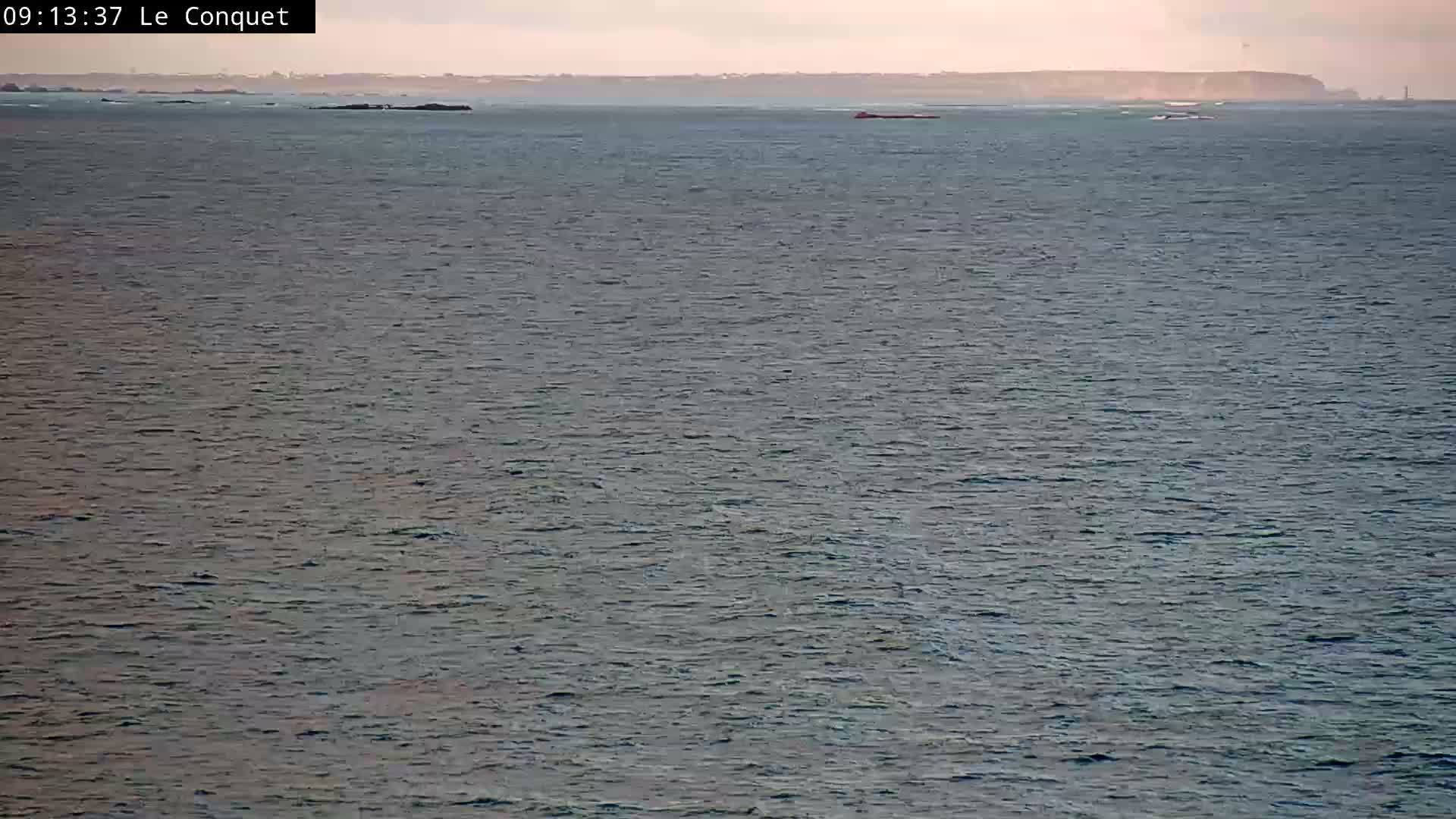 A wide expanse of calm, subtly rippling ocean stretches towards a distant hazy coastline featuring scattered rocky islets, a faint lighthouse, and a small red vessel, all beneath a pale, hazy sky.