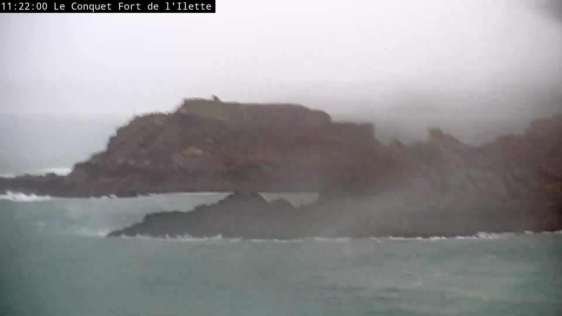 A rocky islet topped with a fort is seen amidst rough, wave-churned waters and smaller rock formations, all under an overcast sky with heavy mist or fog.