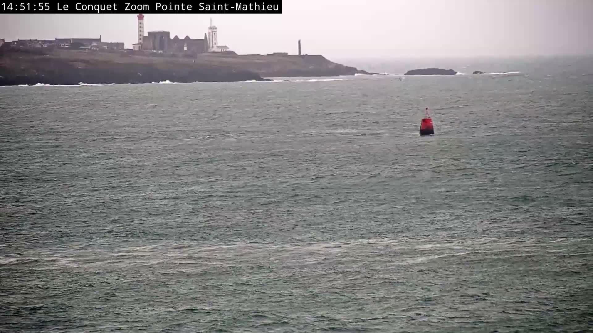 A rocky islet topped with a fort is seen amidst rough, wave-churned waters and smaller rock formations, all under an overcast sky with heavy mist or fog.