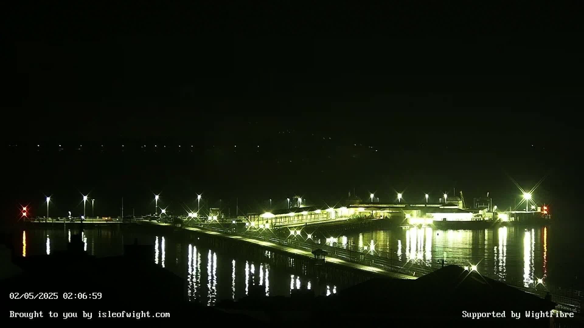 A brightly lit pier and ferry terminal are reflected in the calm, dark water at night.