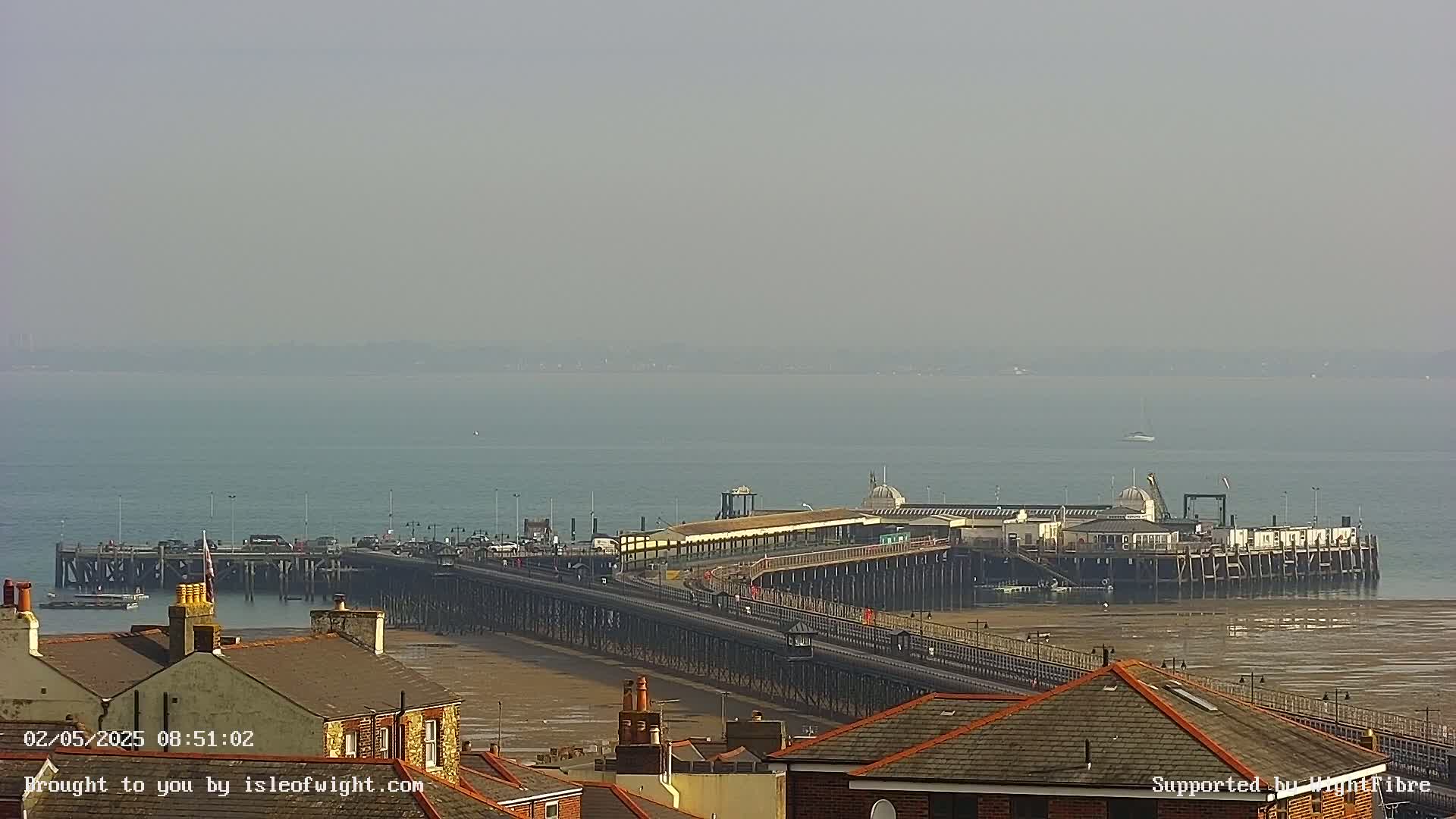A long pier extends over a calm, hazy body of water, with buildings and rooftops visible in the foreground.