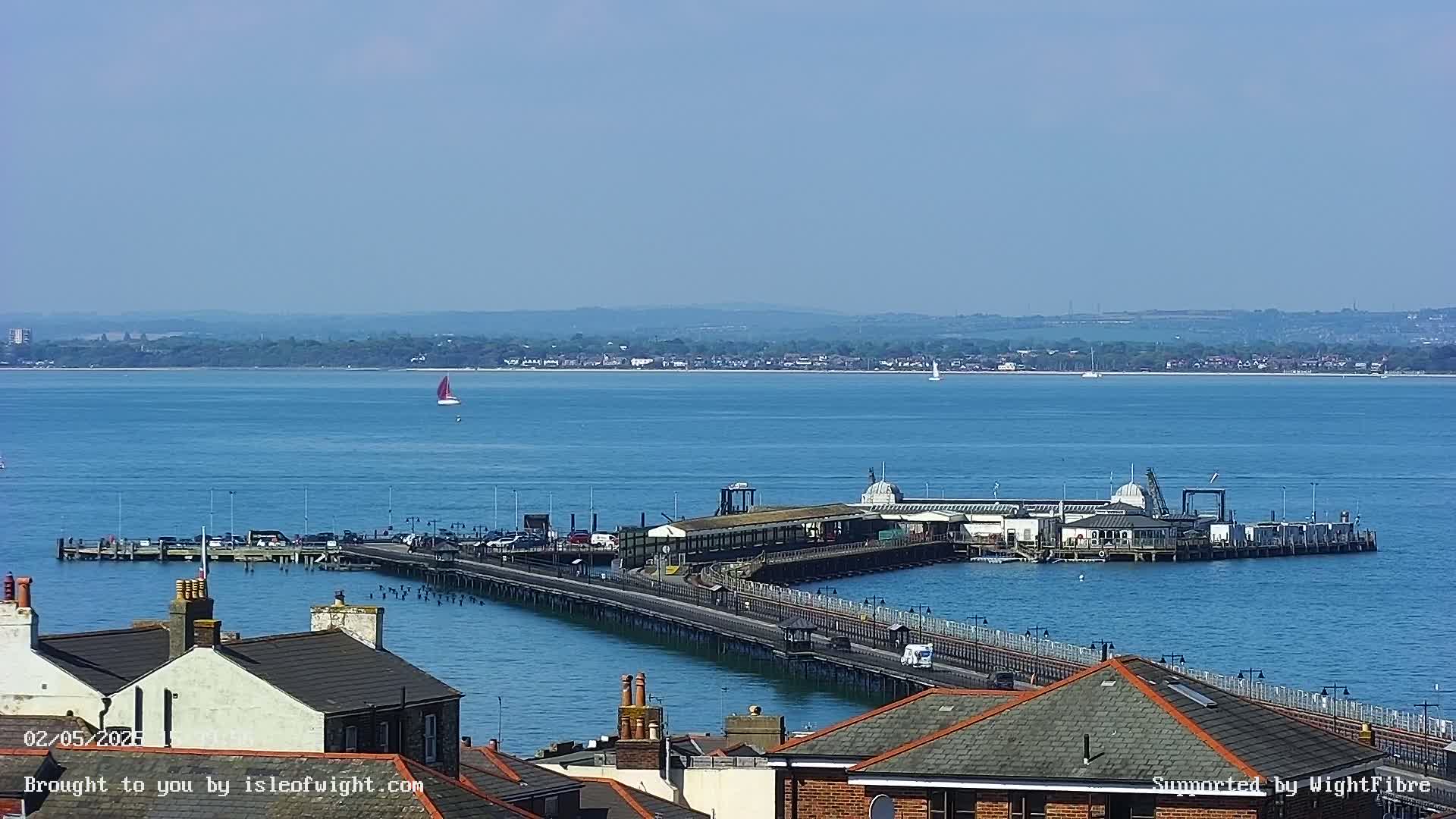 A long pier extends into a calm blue body of water, with a building at its end and rooftops visible in the foreground under a clear sky.