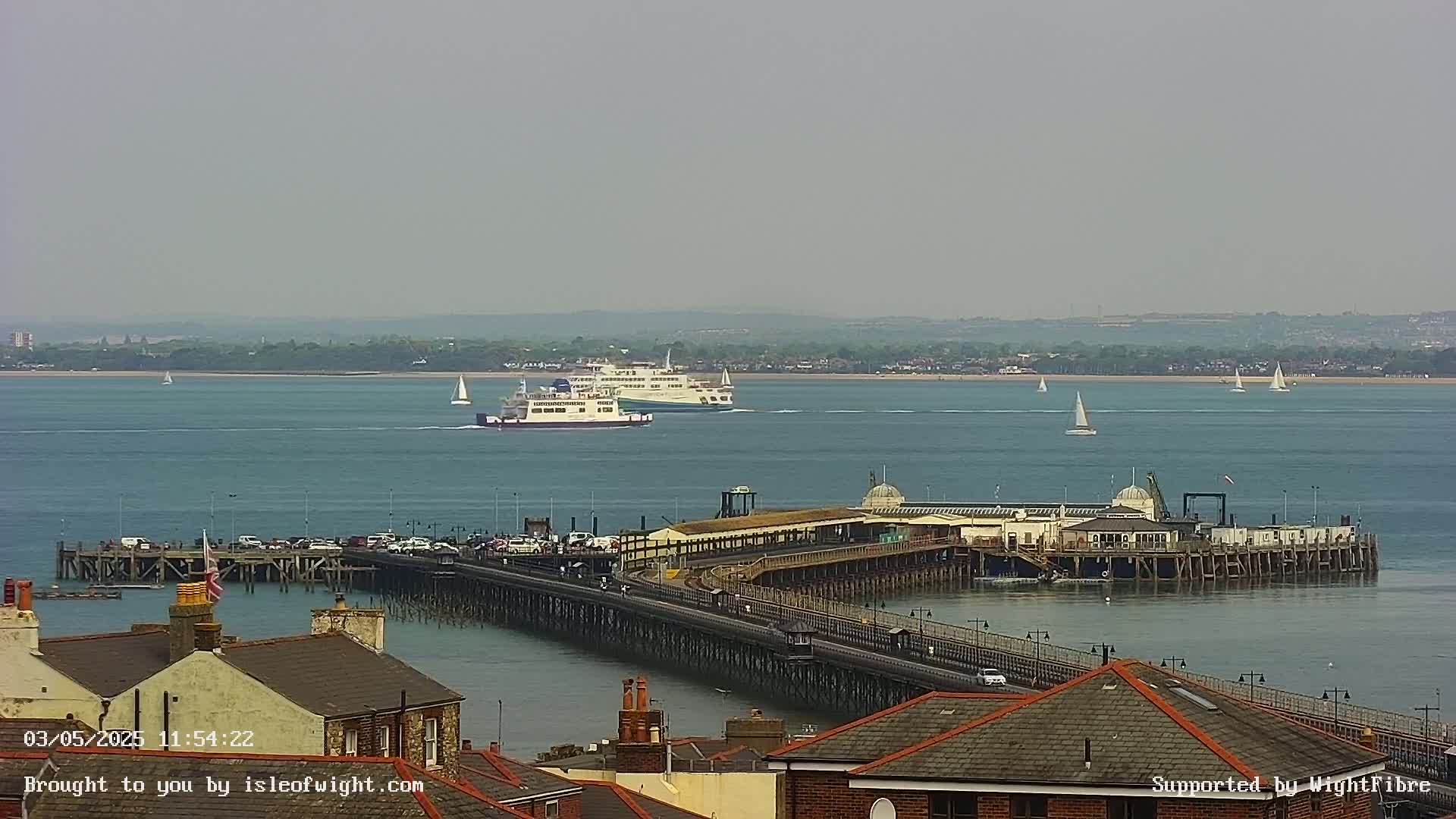 A long pier extends into a calm body of water where two ferries and several sailboats are visible under a hazy sky.