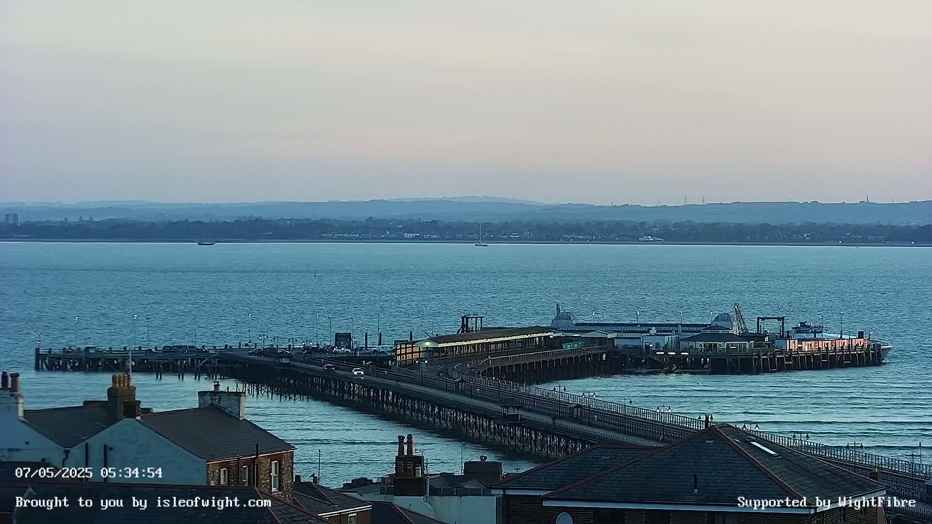 A long pier extends over calm, blue water toward a building complex at the end, with a distant shoreline and hazy sky visible in the background.
