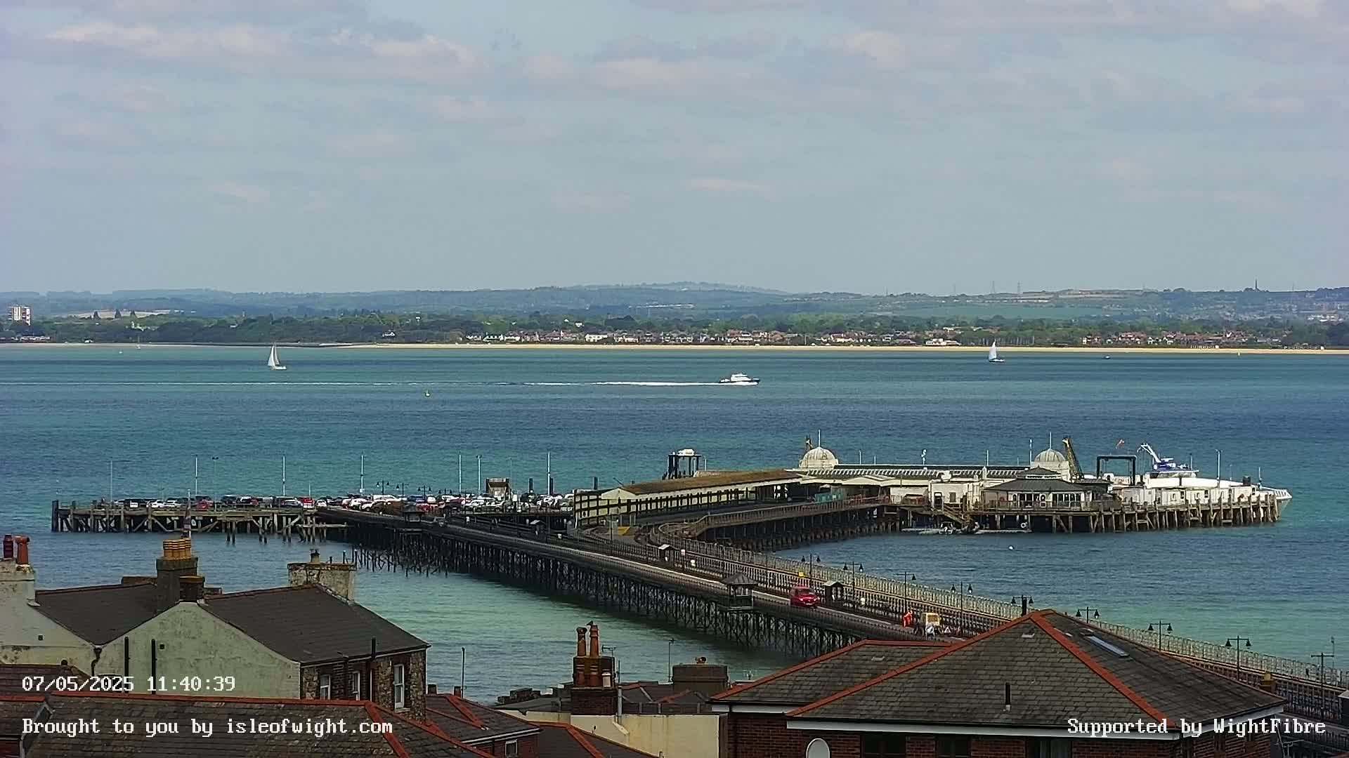 A long pier extends into a calm, blue body of water under a partly cloudy sky, with a distant shoreline and several small boats visible.