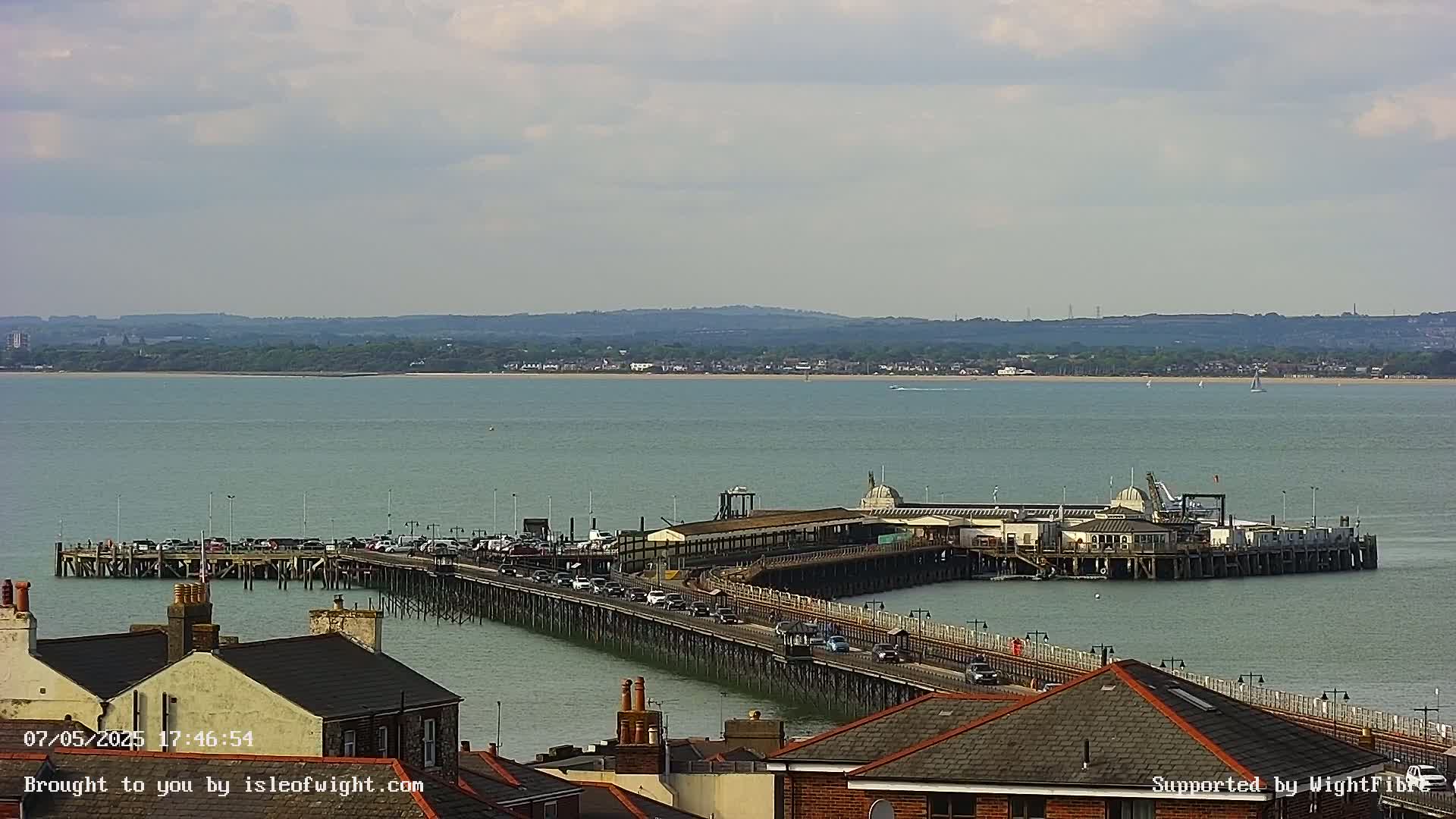 A long pier extends into a calm, light-green sea under a mostly cloudy sky, with cars driving across it and a distant shoreline visible.