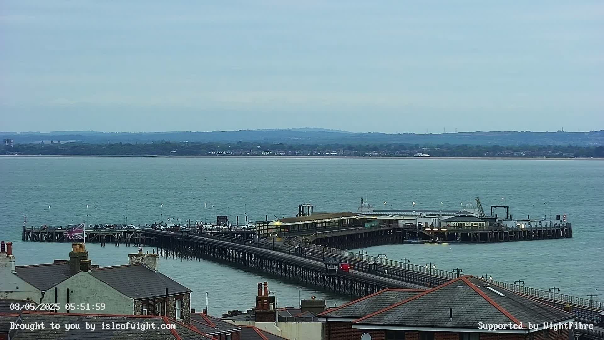 A long pier extends into a calm, blue-green body of water, with a distant shoreline visible under a cloudy sky.
