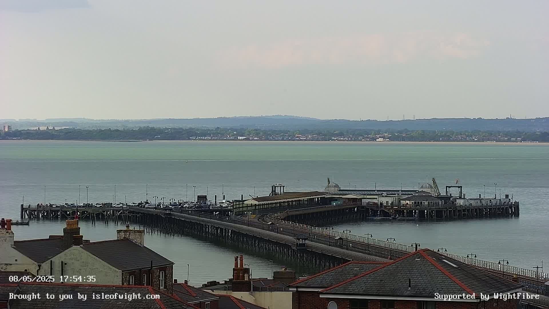 A long pier extends into a calm, light-green body of water, with a distant shoreline visible under a mostly cloudy sky.