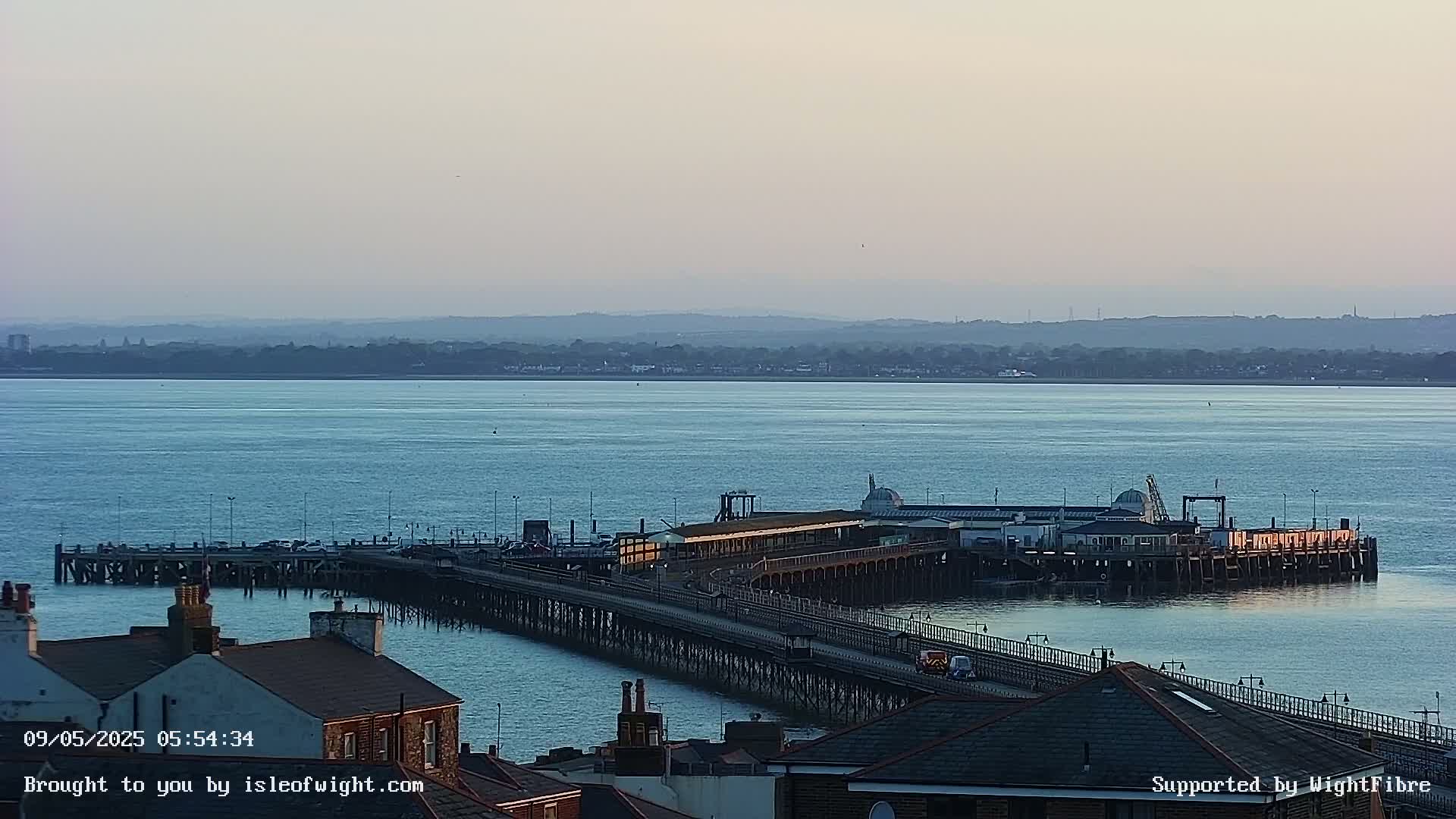A long pier extends from a shoreline into a calm body of water, with a distant landmass visible under a hazy sky.