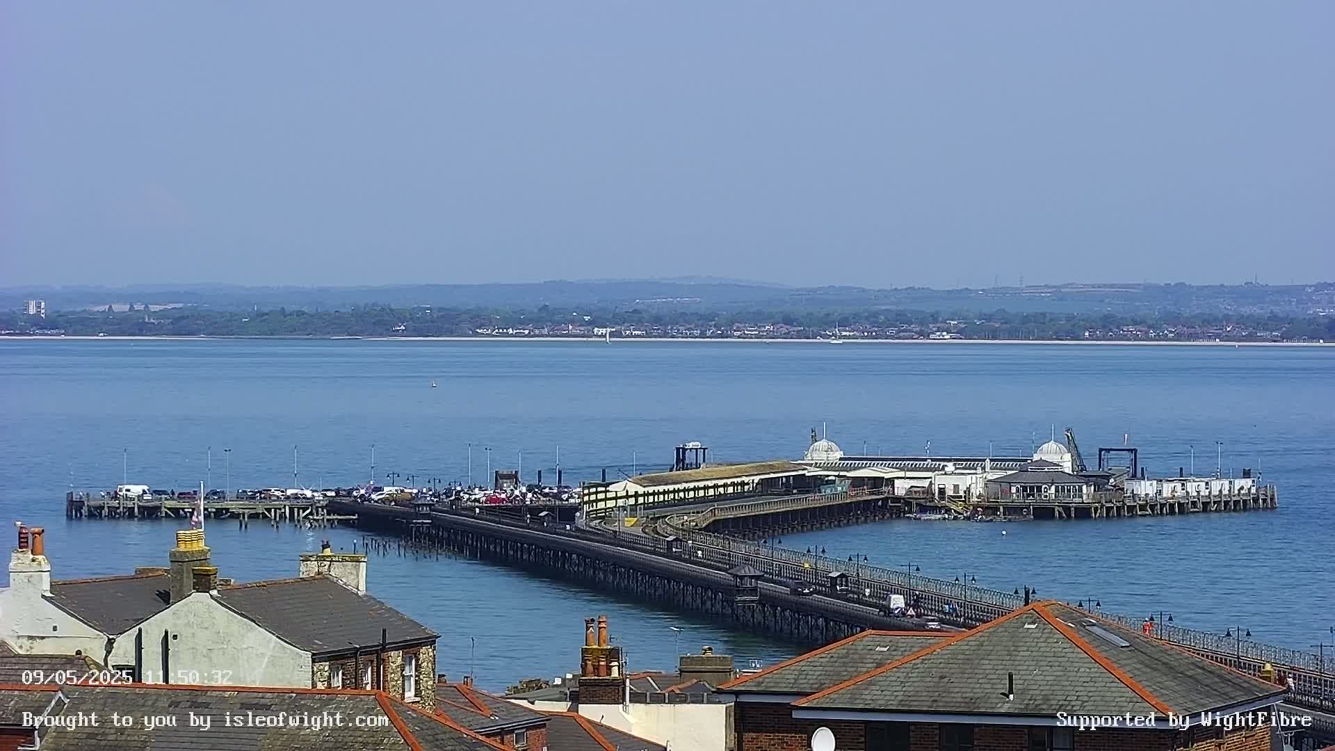 A long pier extends from a shoreline into calm blue water, under a clear sky, with buildings and rooftops in the foreground.